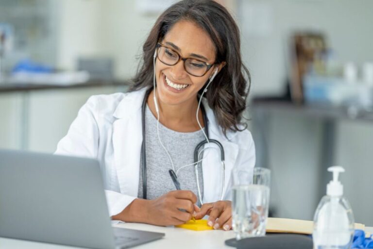 A clinician smiling while taking notes at a laptop during a telehealth visit